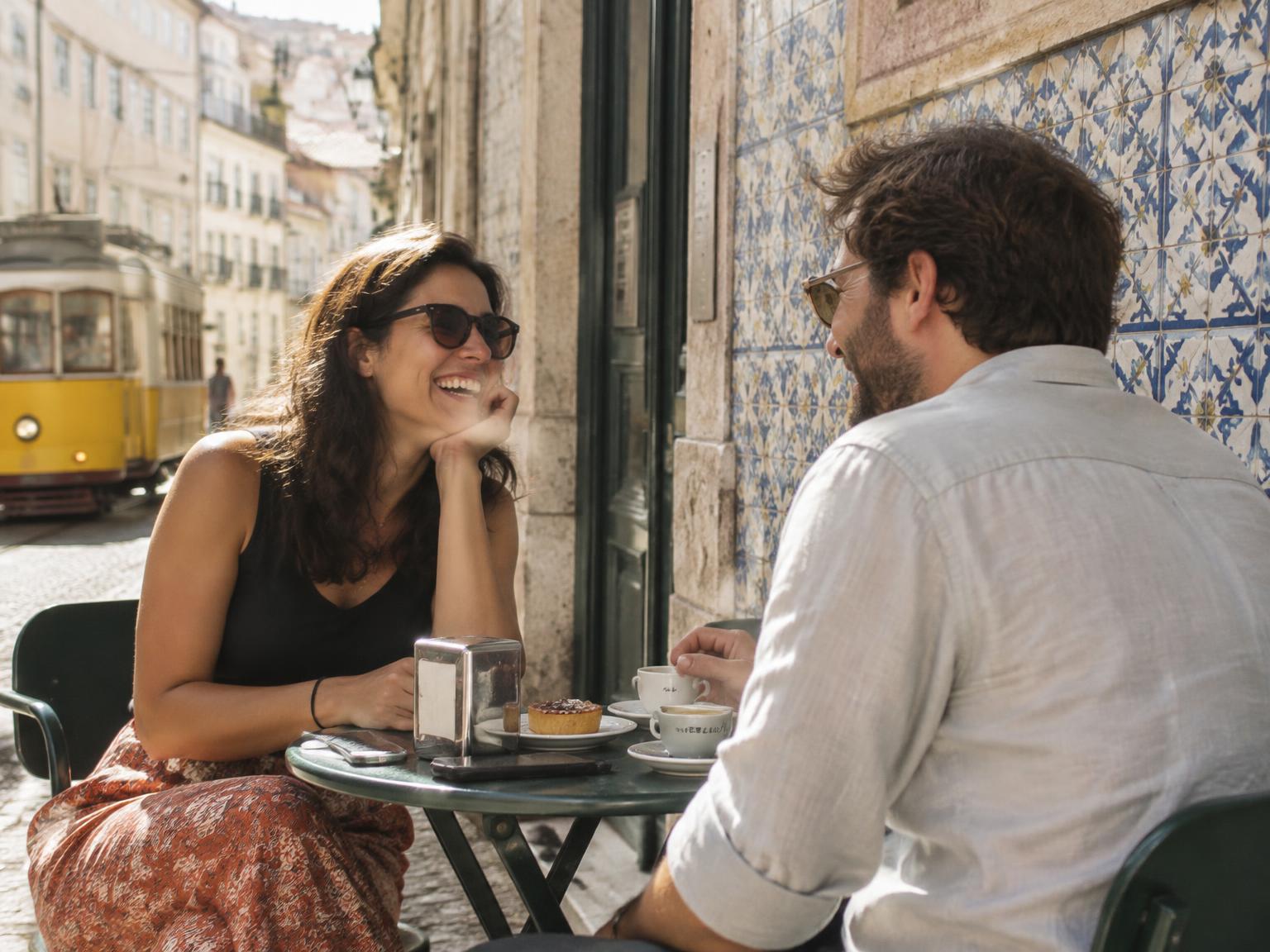 Two friends chatting at a café table on a tiled Lisbon street, with a yellow tram behind them