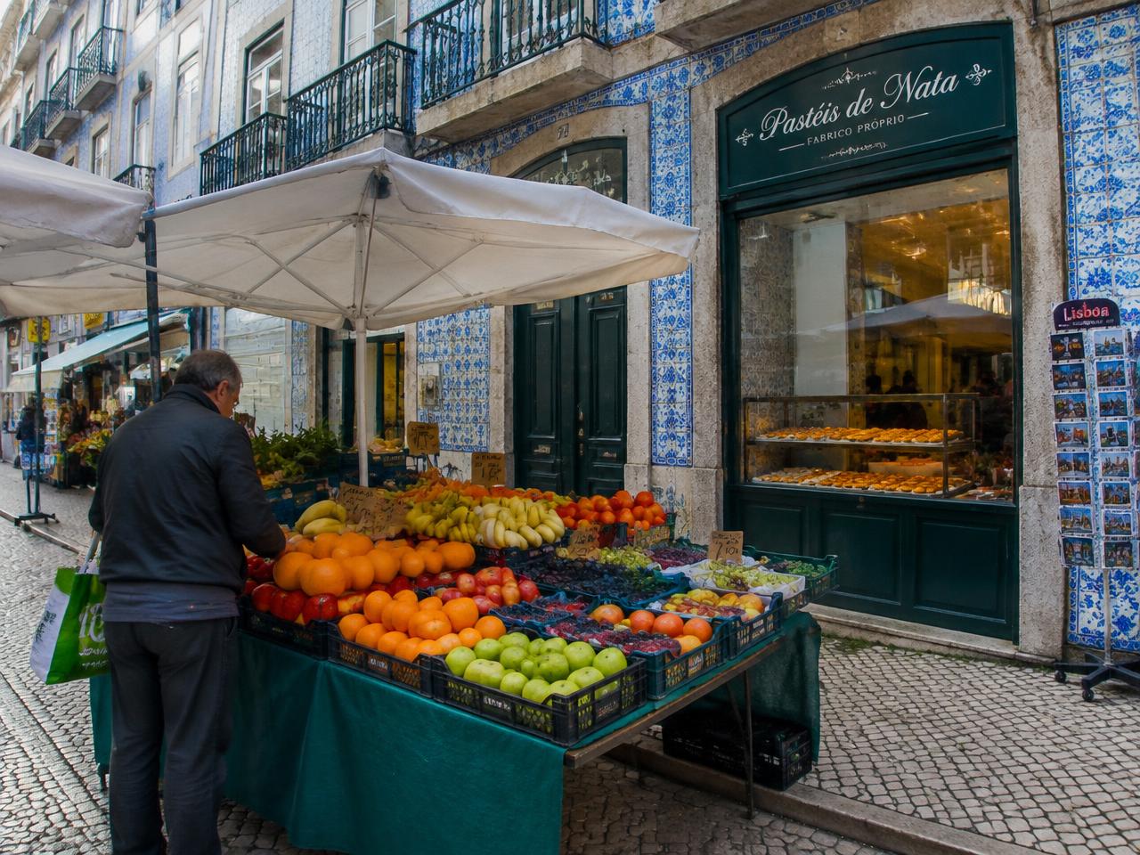 A fruit stall outside a pastéis de nata bakery on a tiled street in Lisbon
